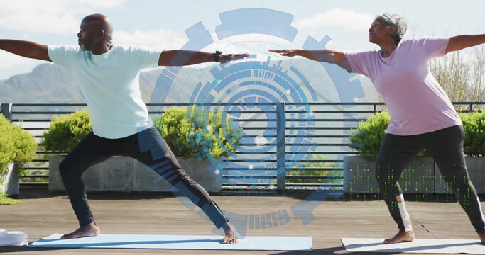 Practicing couple wearing sportswear performing warrior II pose on terrace, with digital interface