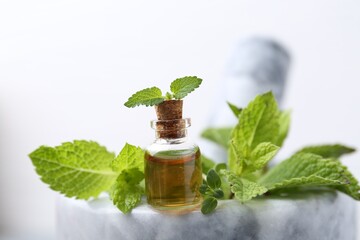 Bottle of aromatic essential oil and herbs in marble mortar against white background, closeup