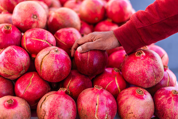 A close-up of a person's hand selecting a vibrant red pomegranate from a neatly arranged fruit display on a checkered table at an outdoor farmers market.