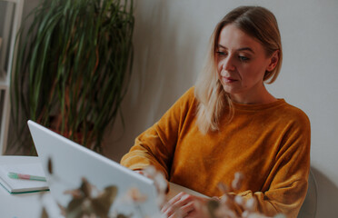 Focused young woman working on a laptop at home. She wears a cozy mustard sweater and sits at a...