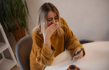 Young tired woman in mustard sweater yawning while holding a smartphone at a table with coffee....