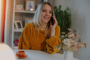 Happy young woman in a mustard sweater having a phone call at a bright table with a small cup and...
