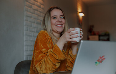 Smiling young woman in a mustard sweater holding a white mug and sitting at a laptop in a cozy home...