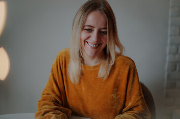 Smiling young woman with blonde hair in a mustard sweater sitting at a white table, looking down...