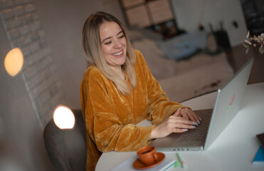 Smiling young woman working on a laptop at home, wearing a mustard sweater and sitting at a round...