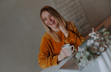 Happy young woman in a mustard sweater laughing while holding a coffee mug, sitting at a desk with...