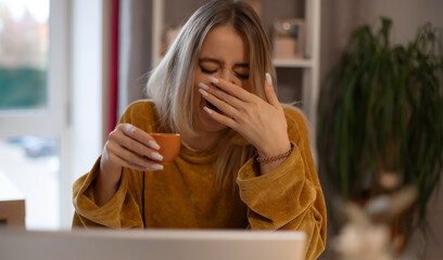 Tired young woman yawning while holding a small coffee cup in a cozy home setting, wearing a...