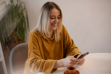 Smiling young woman in a mustard sweater looking at her smartphone while sitting at a table with a...