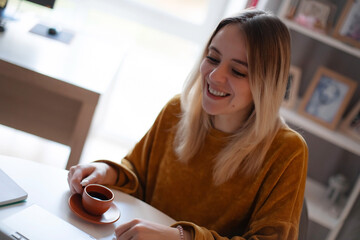 Smiling young woman in mustard sweater enjoying a small cup of coffee while sitting at a desk. Cozy...