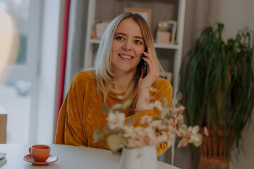 Smiling young woman in mustard sweater having a phone conversation at a cozy table with a coffee...