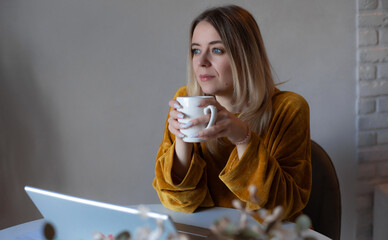 Thoughtful young woman in mustard sweater holding a white mug, looking out while sitting at a desk...