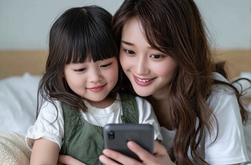 A Japanese woman and child watching a video on a smartphone. The mother, with a ponytail, is smiling at the phone screen while sitting next to the girl, who is holding a pillow in the living room.