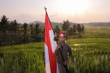 A boy in traditional Indonesian style carrying a red and white flag on a stick in the middle of a rice field with the sun setting in the open air