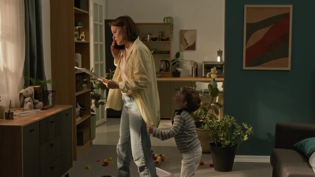 Naughty boy pulling mothers shirt to seek for attention when she speaking on phone holding documents during remote workday