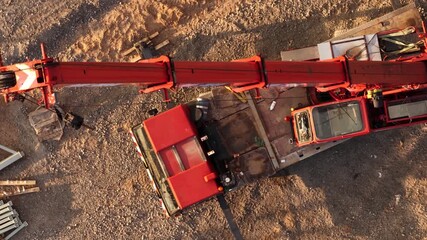 Top-down aerial view of red mobile crane on a construction site, with extended boom, gravel ground, and scattered building materials in warm evening light.