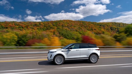 Fototapeta premium White SUV driving through vibrant autumn forest on highway with colorful trees and mountains under a clear blue sky