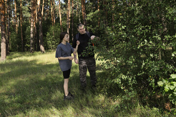 Man teaching teenage girl about edible and wild plants during forest hike, examining leaves and berries, promoting outdoor learning, survival skills, and ecological literacy.