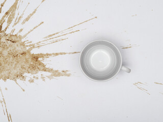 A white coffee cup sits beside a large, splattered coffee stain on a white surface.