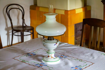 Vintage oil lamp on an embroidered tablecloth, placed on a table in a traditional interior with a tiled stove and wooden chairs