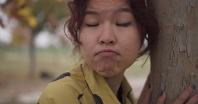 Worried Young Woman Hiding Behind a Tree in an Autumn Park, Making Faces and Puffing Cheeks