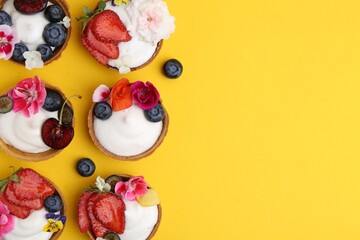 Sweet tartlets with berries and flowers on yellow background, flat lay with space for text. Delicious dessert