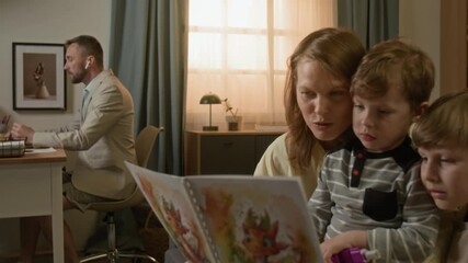 Zoom out rack focus view of young man working remotely on laptop at table while his wife and two kids reading picture book sitting on couch together - Powered by Adobe