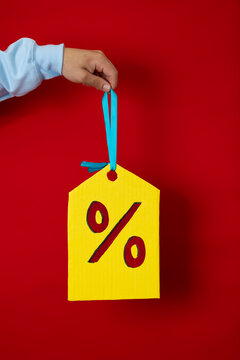 Male hand in a blue sweatshirt holding a huge yellow discount tag with his finger on a red background, concept of sales and good prices