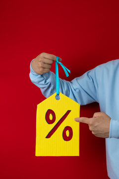 Unrecognizable man in a blue sweatshirt pointing with his hand at a huge yellow discount tag on a red background, good price concept