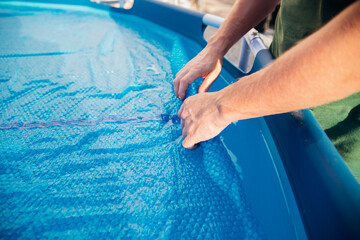 The blue pool cover is assembled into a roll by men's hands, the concept of summer recreation and pool maintenance.