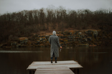 Woman in a gray coat and beanie stands on a wooden pier, holding a wine glass, facing a calm river...