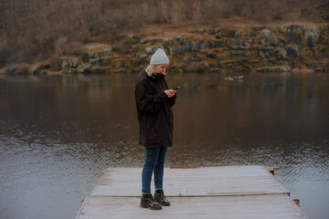 Young woman in a winter jacket and beanie standing on a wooden dock, looking at her phone, surrounded by calm water and forested rocky hills in a moody outdoor setting.