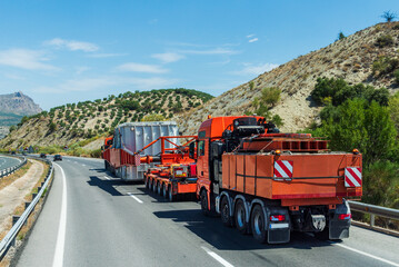 Heavy haul trucks transporting a massive metal crate on a steep incline, with one truck in front and another behind the load. Rear view.