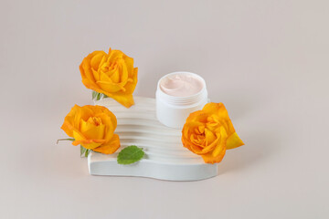 Face cream in a jar on a white wavy podium and yellow rose flowers on a beige background