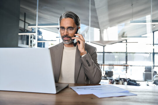 Mature professional businessman executive making call on cell using computer communicating with client. Busy middle aged business man talking on mobile phone working on laptop sitting at office desk. - Powered by Adobe