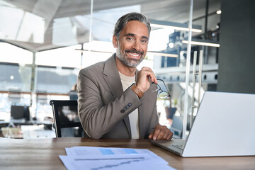 Smiling entrepreneur wearing suit sitting at desk in office looking at camera, portrait. Happy...