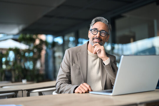 Smiling busy middle aged professional male entrepreneur sitting at table outdoors hybrid working. Happy mature business man entrepreneur using laptop computer outside office looking away. Copy space