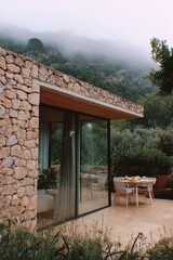 Obraz premium A contemporary stone house gracefully situated amidst the enchanting, misty mountains, featuring an inviting outdoor dining area beautifully arranged for breakfast, basking in the gentle morning light