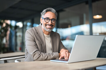 Portrait of happy confident mature professional business man working looking at camera. Smiling middle aged businessman wearing glasses sitting at outdoor office table using laptop. Copy space.