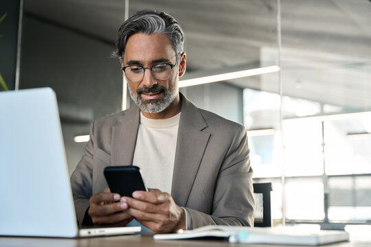 Smiling middle aged business man manager sitting in office using mobile cell phone. Mature businessman professional executive holding cellphone working at desk with laptop and smartphone.