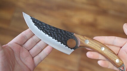 Handcrafted knife with textured dark blade and wooden handle, held over a wooden surface,...
