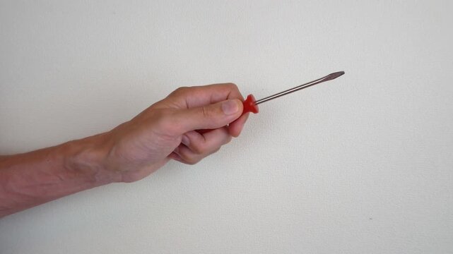 A man's hand holds a screwdriver with a red handle, pointing the tip to the right. A light wall is visible in the background, a close-up image without additional objects.