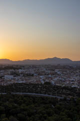 A stunning sunset view over Alicante, showcasing the cityscape and mountains in the background. Trees and roads offer an insight into the surrounding natural beauty.