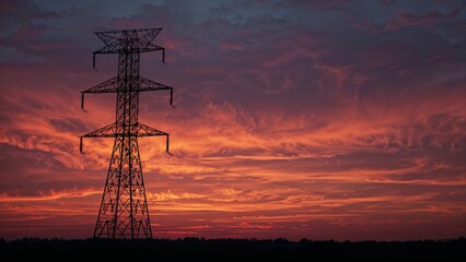 Fototapeta premium High-voltage power transmission tower silhouetted against a sunset, power lines in the background
