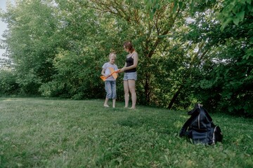 A mother and her son with hearing aids joyfully launch a paper airplane in a sunlit park, capturing a heartwarming moment of inclusive play and bonding in nature