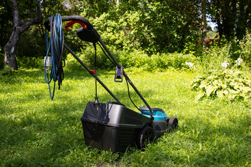 The electric lawn mower stands on the green grass. the power cord is twisted into a spiral. © Светлана Высокос