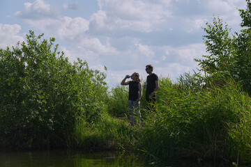 Two individuals converse by a riverbank under a bright sky with scattered clouds during the late...