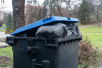 A black trash bin is filled with black garbage bags, located in a park with bare trees and a hint...