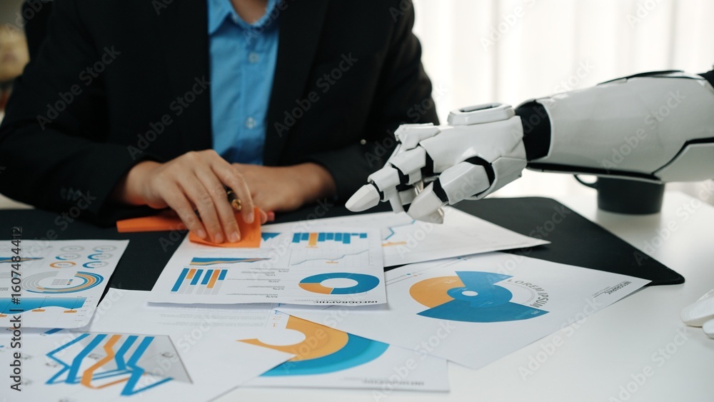 Poster a professional collaborates with a robotic arm, examining financial graphs and charts on a desk, sho
