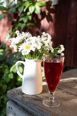 A glass with a red drink on a wooden background, a jug with daisies and gypsophila, a wooden wall...