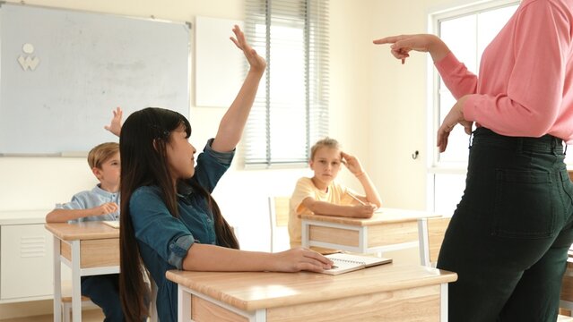 Happy asian student raising hand to volunteer for answering question at elementary class while teacher choosing student to write answer on blackboard. Diverse children attend in classroom. Pedagogy.